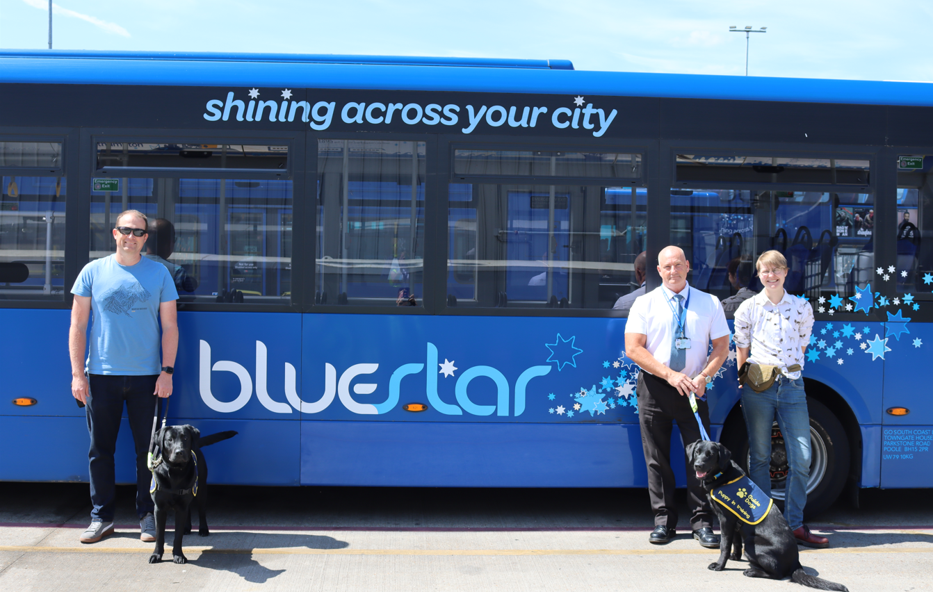 Two Guide Dog puppies next to a Bluestar bus with their trainers